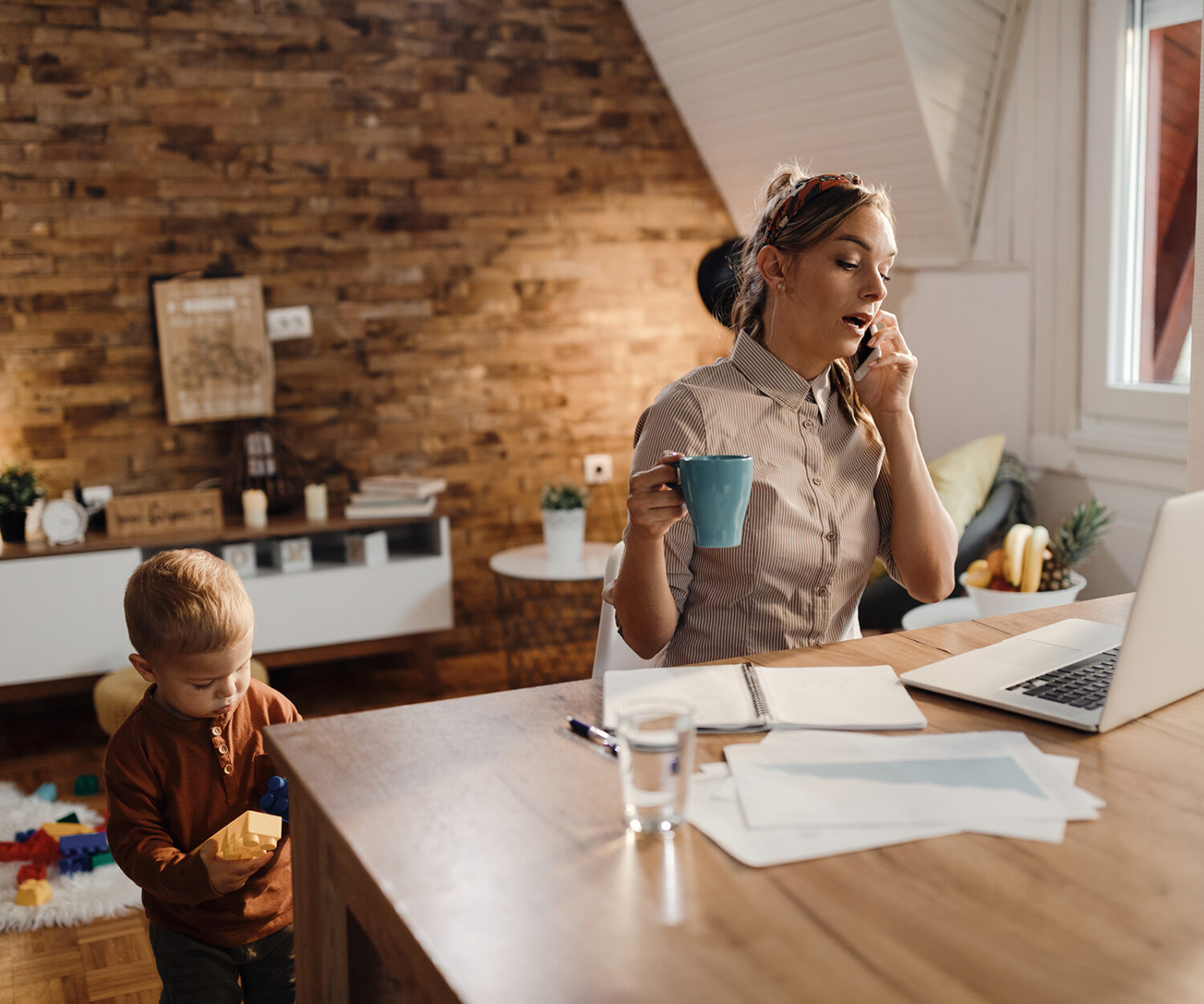 Freelance worker using laptop and communicating over cell phone while being with her son at home.